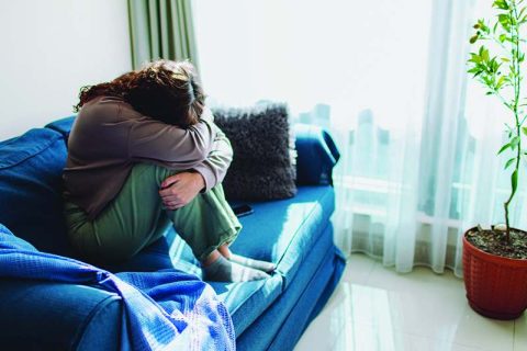 A dismayed caregiver experiencing caregiver PTSD sits on a blue couch upright in the fetal position.