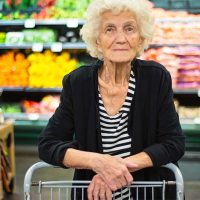 An older adult stands in the produce section of a grocery store, resting her arms on the shopping cart.