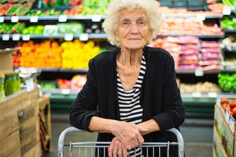 An older adult stands in the produce section of a grocery store, resting her arms on the shopping cart.