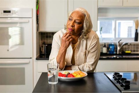 An older adult leans on her counter with a plate of uneaten food in front of her, perhaps indicating a senior with an eating disorder.
