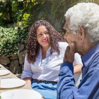 A woman talks to an older adult about the symptoms of dementia outside at a dinner table.