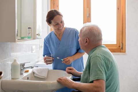 A caregiver providing personal care services in the Syracuse area helps a senior man brush his teeth.